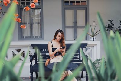 Young woman using smart phone while sitting on chair against house
