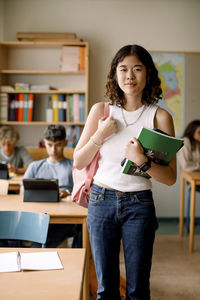 Portrait of teenage girl with book and backpack in classroom