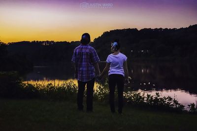 Rear view of couple standing by lake against sky during sunset