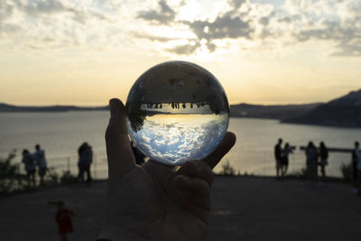 Close-up of hand holding crystal ball by sea against sky during sunset