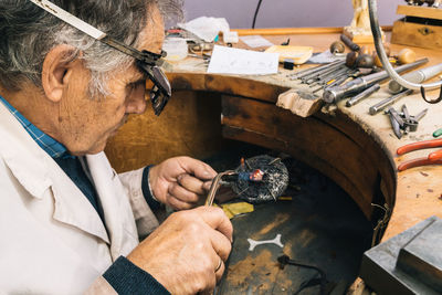 High angle side view of skilled mature male master in glasses using soldering iron while working at workbench with professional instruments in jewelry workshop