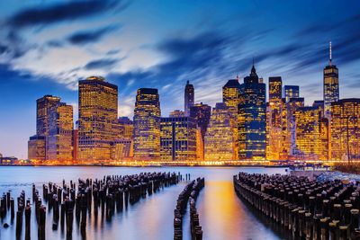 Illuminated buildings by river against sky in city at dusk