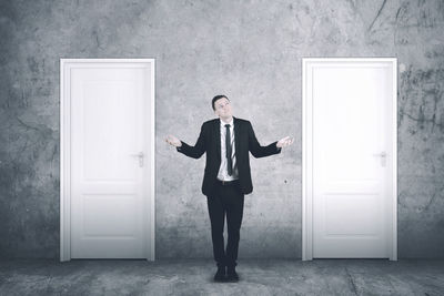 Full length of confused businessman standing by closed doors against wall