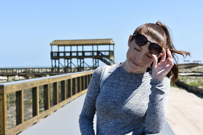 Smiling young woman with eyes closed standing by railing against clear sky