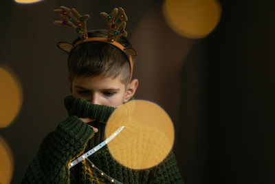 Cute boy in deer horns and a knitted sweater hides his face in collar as in a protective mask.