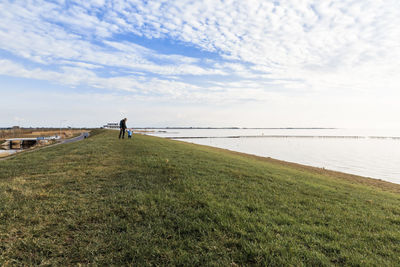 Father and son walking on field by lake against sky