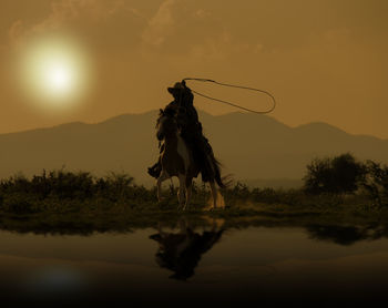 Cowboy riding horse by lake against sky during sunset