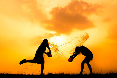 Low angle view of silhouette siblings playing with water against orange sky