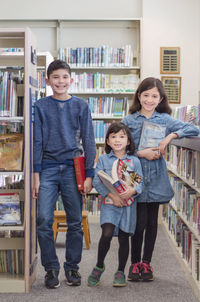 Full length portrait of cute smiling friends holding books while standing in library