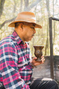 Side view of argentinian male drinking mate from calabash gourd near kettle against rack above burning fire in countryside