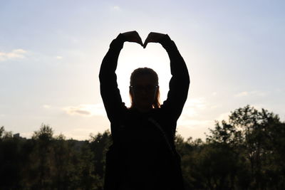 Woman making heart shape with hands while standing against sky during sunset