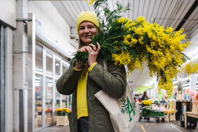 Woman in yellow cap and large bouquet of mimosa flowers at flower market