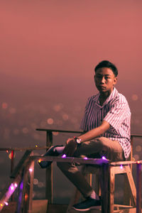 Young man sitting on chair against cityscape during sunset