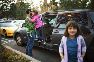 Man assisting daughter in disembarking from car with children in foreground