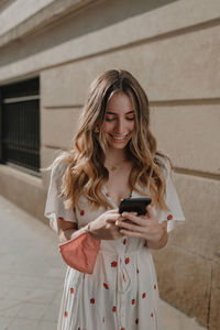 Beautiful young woman using phone while standing against wall