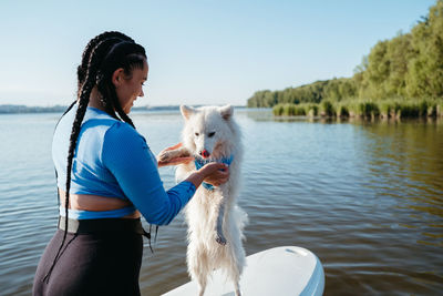 Young woman with locs holding her pet japanese spitz on the lake, dog standing on the sup board