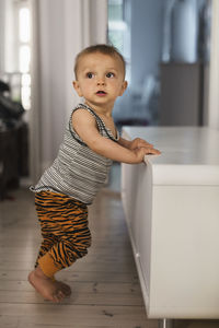 Full length of baby boy leaning on sideboard at home