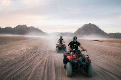 Friends riding quadbikes at desert against sky during sunset