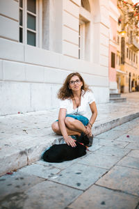 Full length portrait of woman stroking cat on footpath in city