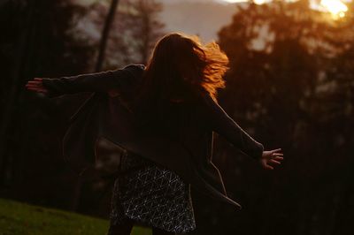 Rear view of cheerful woman with arms outstretched dancing at park during sunset