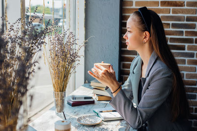 A young woman is sitting at a table by the window in a cafe and drinking coffee.