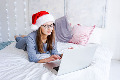 Pretty woman sitting on bed working with laptop wearing santa claus hat