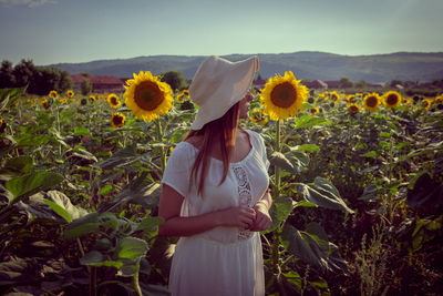 Teenage girl wearing hat standing amidst sunflowers against clear sky