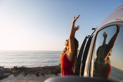 Young woman with hand raised standing by camper van at beach