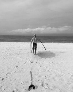 Rear view of man carrying paddleboard towards sea against cloudy sky