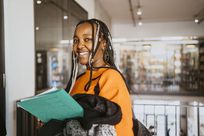 Portrait of smiling braided female student holding jacket and file in university