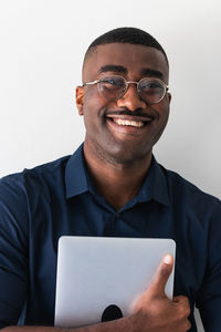 Positive african american adult male in formal clothes and eyeglasses standing with netbook in hands and looking at camera on white background