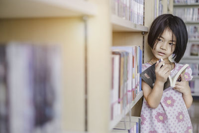 Girl looking away while holding book by shelves in library