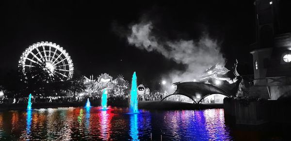 Illuminated ferris wheel by river against sky at night