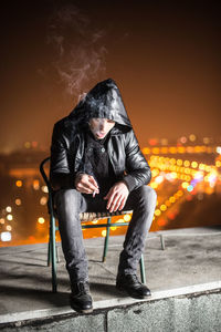 Full length of man smoking cigarette while sitting on chair at building terrace during night