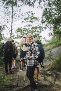 Side view of smiling senior woman holding pole during hiking with friends in forest