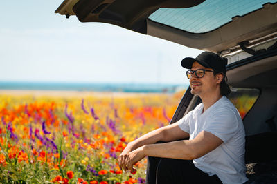 Young man wearing sunglasses traveling on suv car trunk amidst colorful flowering plants in summer