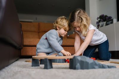 Cute little curly haired siblings sitting on floor and playing with toy road and cars while spending time together in living room at home