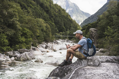 Mature man using smart phone sitting on rock by river