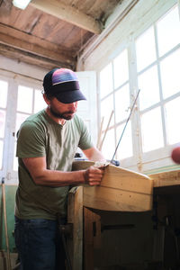 Concentrated bearded male worker in cap and t shirt making longboard at workbench in daylight