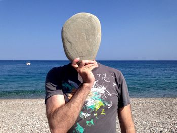 Man covering his face with stone at beach against clear blue sky during sunny day