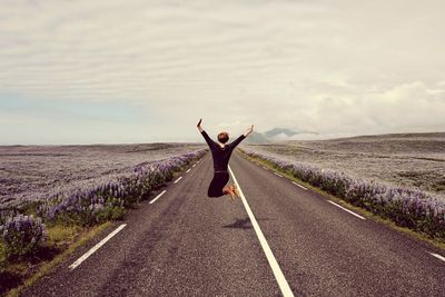 Woman jumping on road amidst flowering field against sky