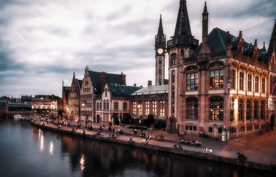 People in front of buildings by river against cloudy sky