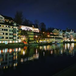 Illuminated buildings by lake against sky at night