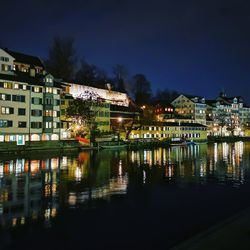 Illuminated buildings by lake against sky at night