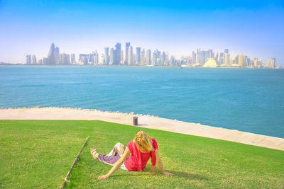 Woman sitting on lawn against sea in city