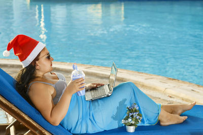 Side view of woman wearing santa hat using laptop by swimming pool