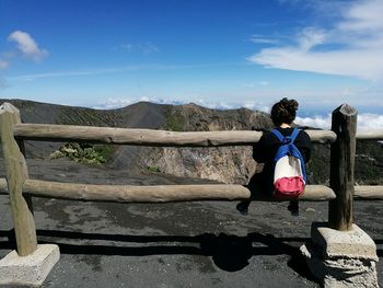 Rear view of young woman sitting on fence against mountains