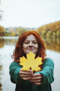 Redhead funny woman walking with yellow leaf near lake in autumn park.