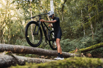 Low angle of fit sportsman in helmet and cycling goggles carrying bicycle on shoulder while walking on fallen tree trunk in woods