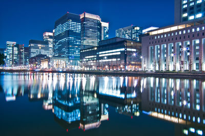 Illuminated modern buildings by river against sky at night